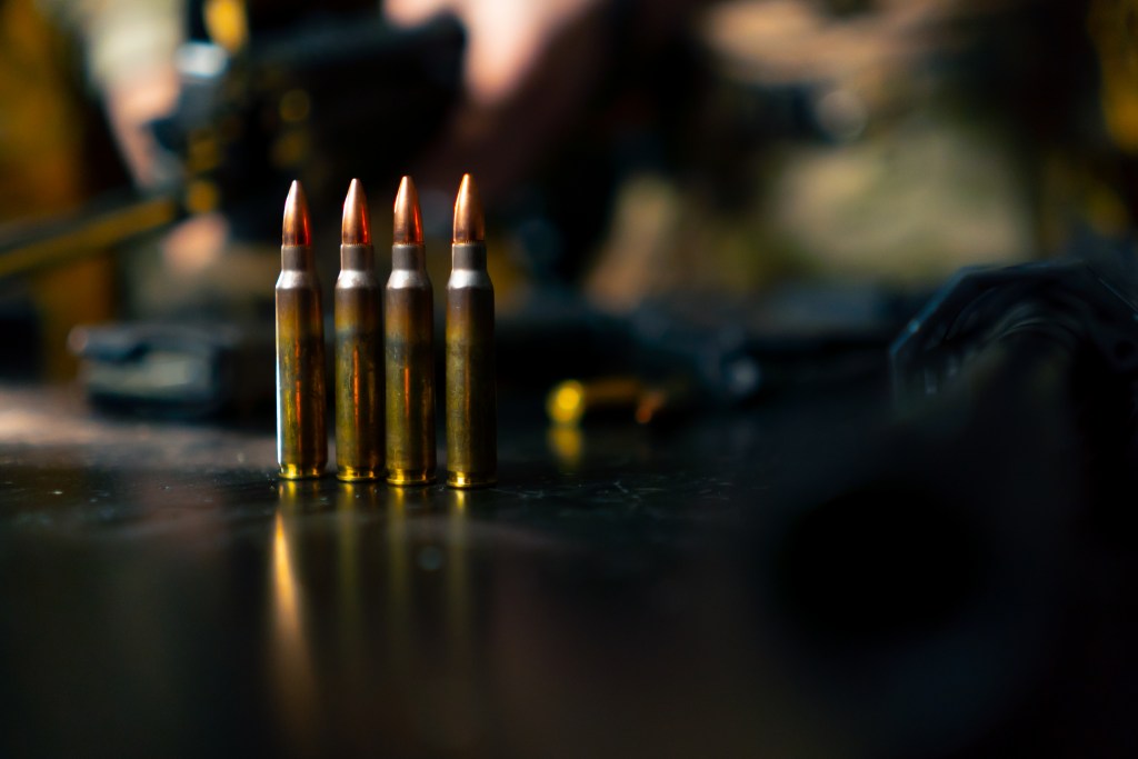 rifle cartridges standing upright on a black surface with black firearm magazines and parts blurred in the background.
